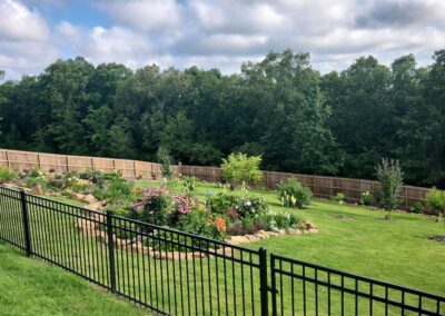 A well-maintained backyard garden with a variety of plants and flowers, bordered by a wooden fence and a metal railing. - Little Rock Lawn Care and Mowing Services by Apex Lawn Care Lush garden landscape with vibrant flowers and greenery, surrounded by a black metal fence, showcasing professional lawn care services by APEX Lawn Care in Little Rock, Arkansas.