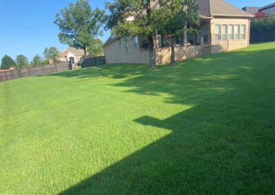 A well-maintained lawn in front of a suburban house with a clear shadow of an employee on the grass. - Little Rock Lawn Care and Mowing Services by Apex Lawn Care Lush green lawn maintained by APEX Lawn Care, showcasing professional lawn care services in Little Rock, Arkansas, with a well-kept residential yard and clear blue sky.