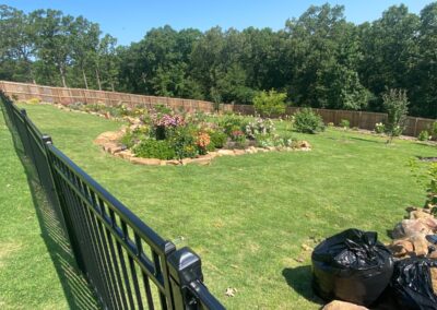 A well-manicured backyard garden with a variety of flowers and plants, surrounded by a fence, with a row of filled black trash bags in the foreground provides an engaging workspace for any employee. - Little Rock Lawn Care and Mowing Services by Apex Lawn Care Lush green lawn featuring a circular flower garden surrounded by a decorative stone border, with a black fence in the foreground, showcasing APEX Lawn Care's landscaping services in Little Rock, Arkansas.