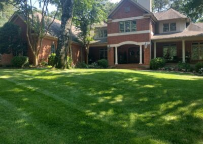 A two-story brick house with large windows, a prominent chimney, and a well-manicured front lawn with a shady tree. - Little Rock Lawn Care and Mowing Services by Apex Lawn Care Lush green lawn with neatly mowed stripes in front of a well-maintained brick home, showcasing professional lawn care services by APEX Lawn Care in Little Rock, Arkansas.