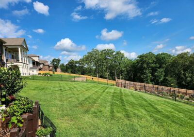 An employee meticulously maintains a lawn next to a residential area in the Little Rock Metro Area, all secured with a metal fence under a clear blue sky. - Little Rock Lawn Care and Mowing Services by Apex Lawn Care Lush green lawn in a residential area of Little Rock, Arkansas, showcasing professional lawn care services by APEX Lawn Care, with a clear blue sky and trees in the background.
