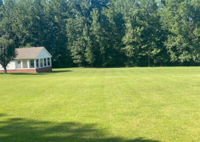 An employee in the response with the word employee and the Little Rock Metro Area a small white building with a red door adjacent to a mowed lawn and a line of trees in the background on a sunny day. - Little Rock Lawn Care and Mowing Services by Apex Lawn Care Lush green lawn maintained by APEX Lawn Care, featuring a well-kept yard with a house and surrounding trees in Central Arkansas.