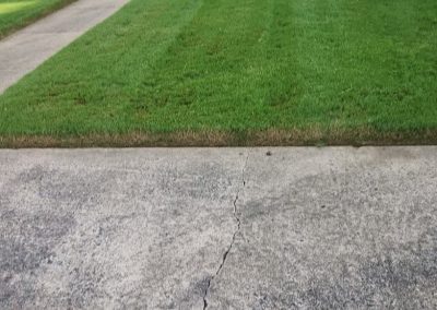 large-17 Lush green lawn with neatly trimmed grass adjacent to a cracked concrete driveway, showcasing APEX Lawn Care's professional lawn maintenance services in Central Arkansas.
